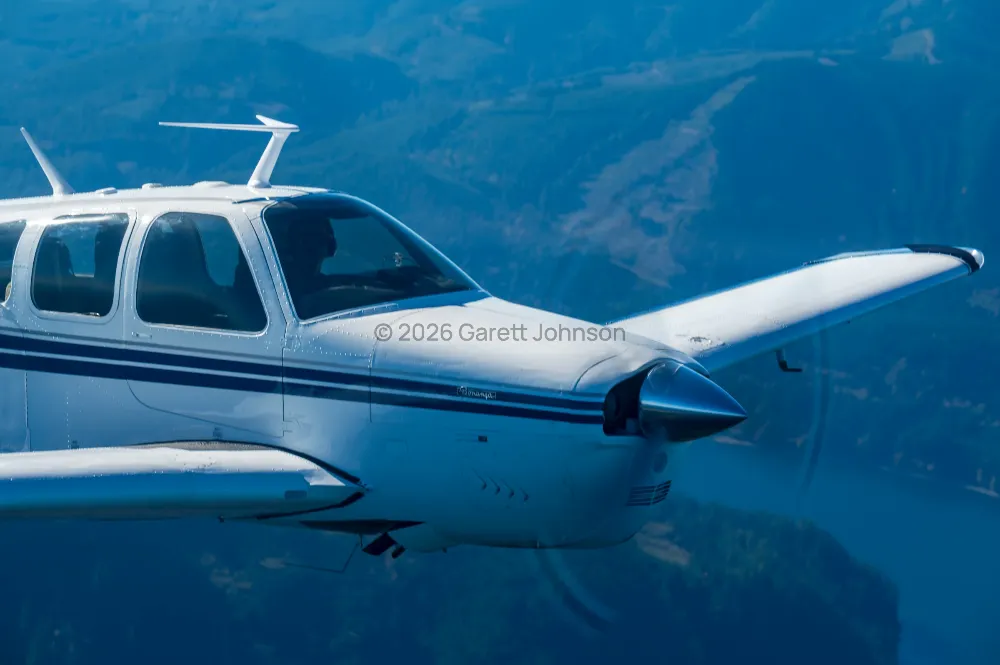 White V-tail Beechcraft Bonanza with navy stripes banks in flight over blue, forested hills and a river, its propeller blurred.