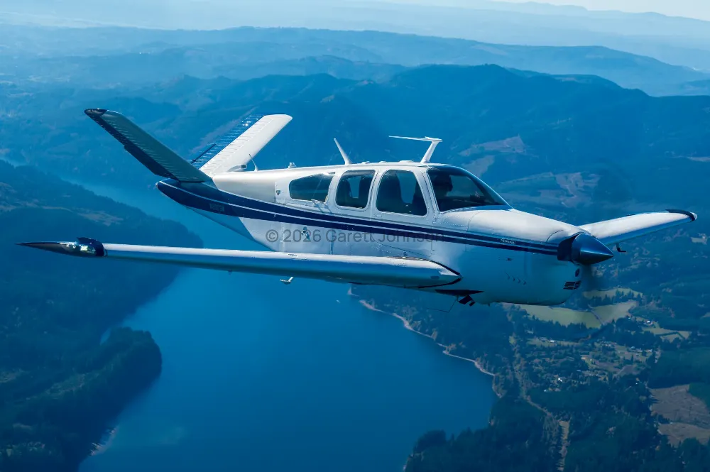 White V-tail Beechcraft Bonanza with dark blue stripes banking over a blue lake and forested hills in hazy sunlight.