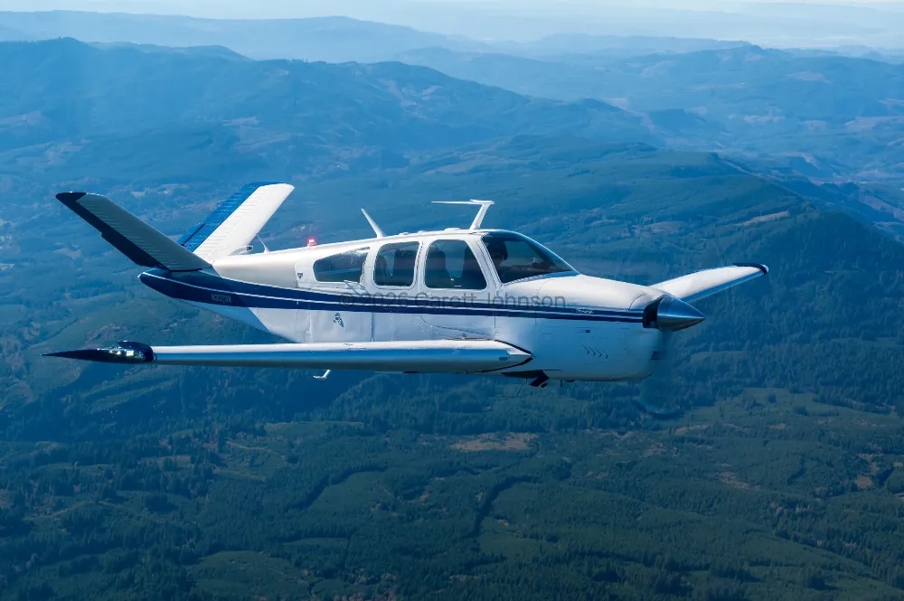 White V-tail Beechcraft Bonanza with navy stripes banks in flight over forested, hazy blue mountain ridges in an air-to-air shot.
