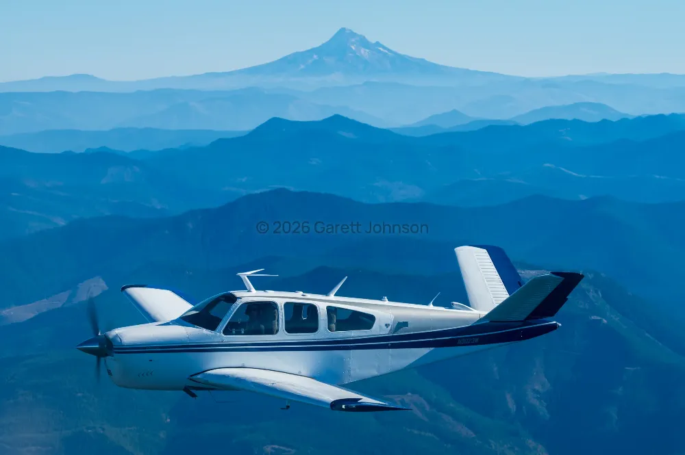 White V-tail Beechcraft Bonanza with dark blue stripes banks in flight over layered blue mountain ridges and a distant snowy peak.