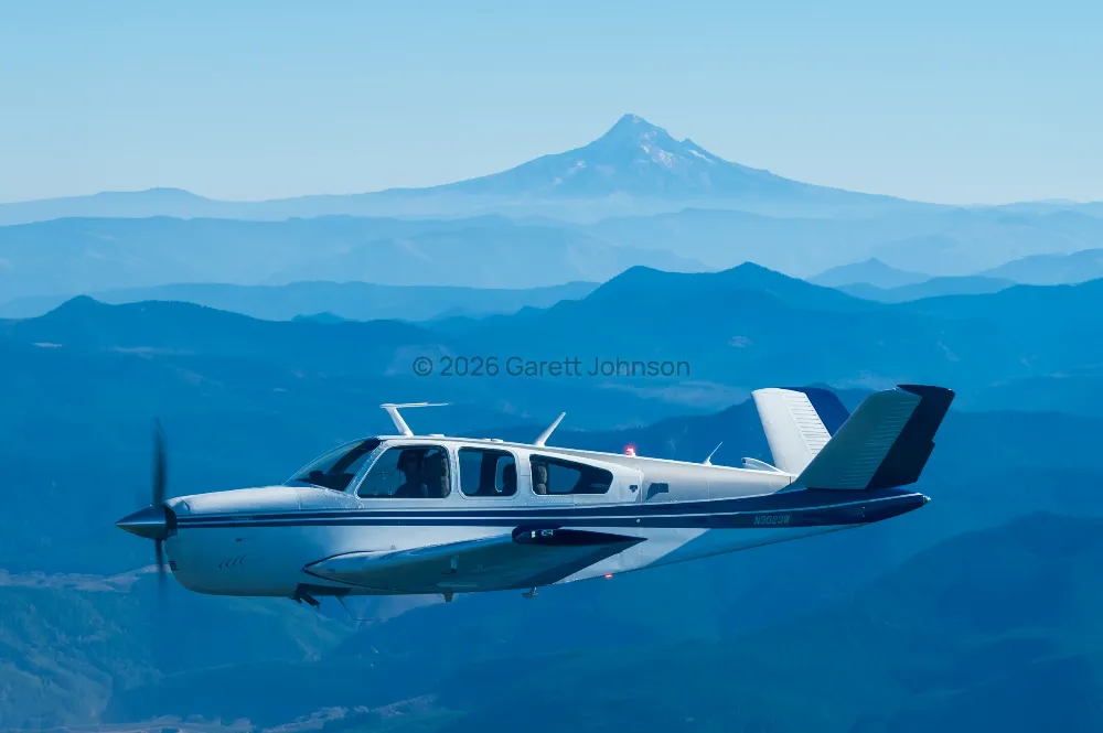 V-tail Beechcraft Bonanza in flight over layered blue ridgelines, with a snow-capped volcanic peak on the hazy horizon.
