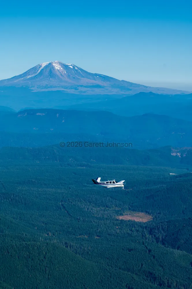 V-tail Beechcraft Bonanza flying over dense evergreen forest, with a snow-dusted volcanic peak rising in the hazy blue distance.
