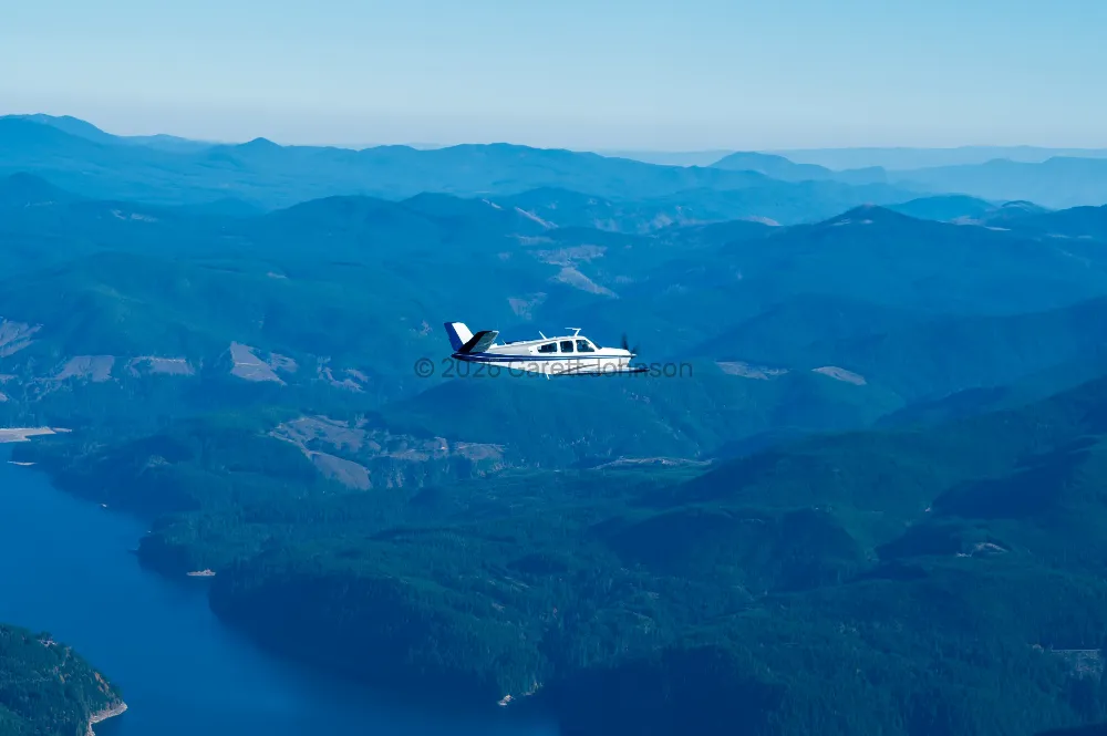White and blue V-tail Beechcraft Bonanza in flight over forested green mountains and a deep blue reservoir under a hazy sky.