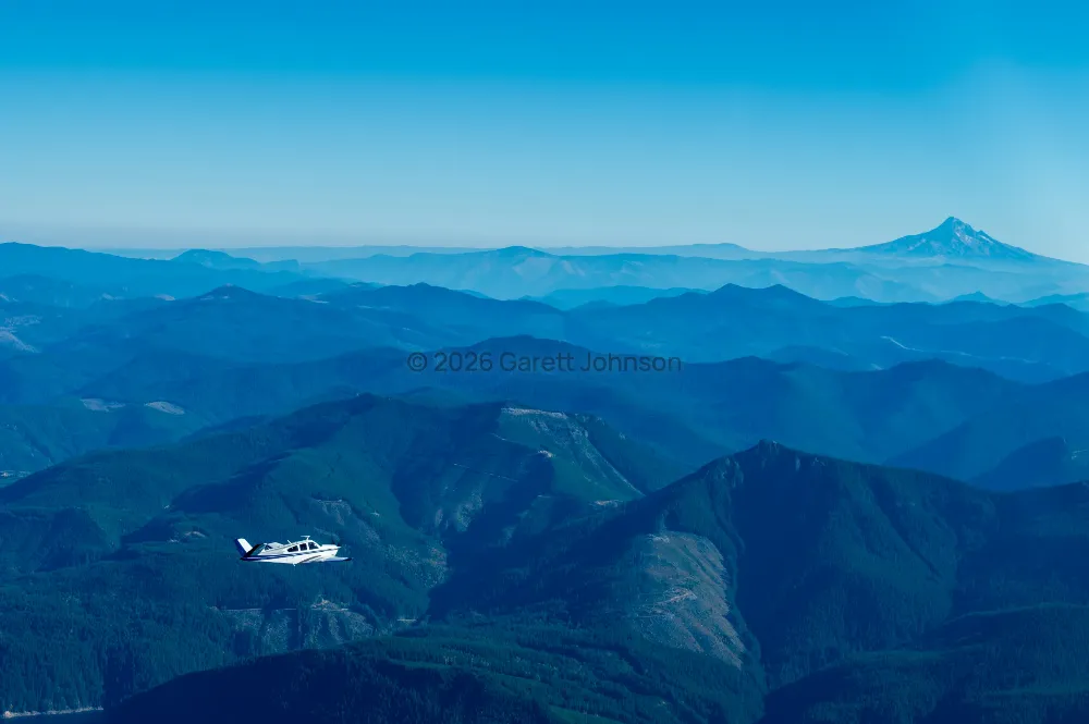 A V-tail Beechcraft Bonanza flies over layered, forested blue ridgelines, with a snow-capped peak on the distant horizon.