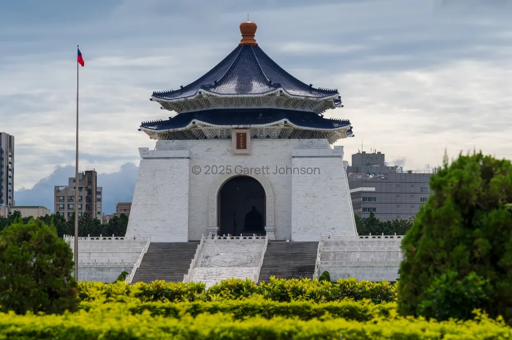 Chiang Kai-shek Memorial Hall in Taipei, a white building with a blue octagonal roof, framed by green hedges under a cloudy sky.