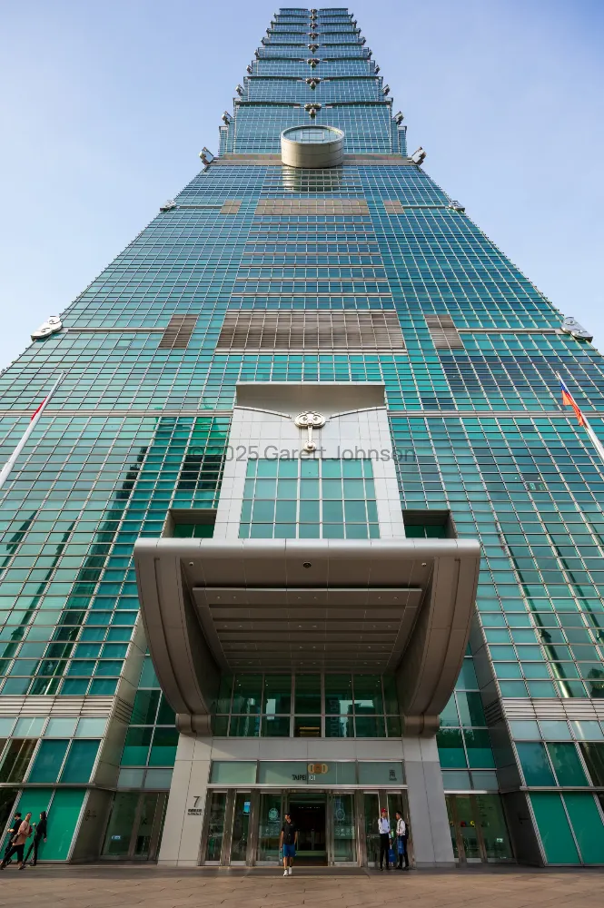 Looking straight up at Taipei 101's green glass facade and tiered tower from its entrance, where a few people stand below.
