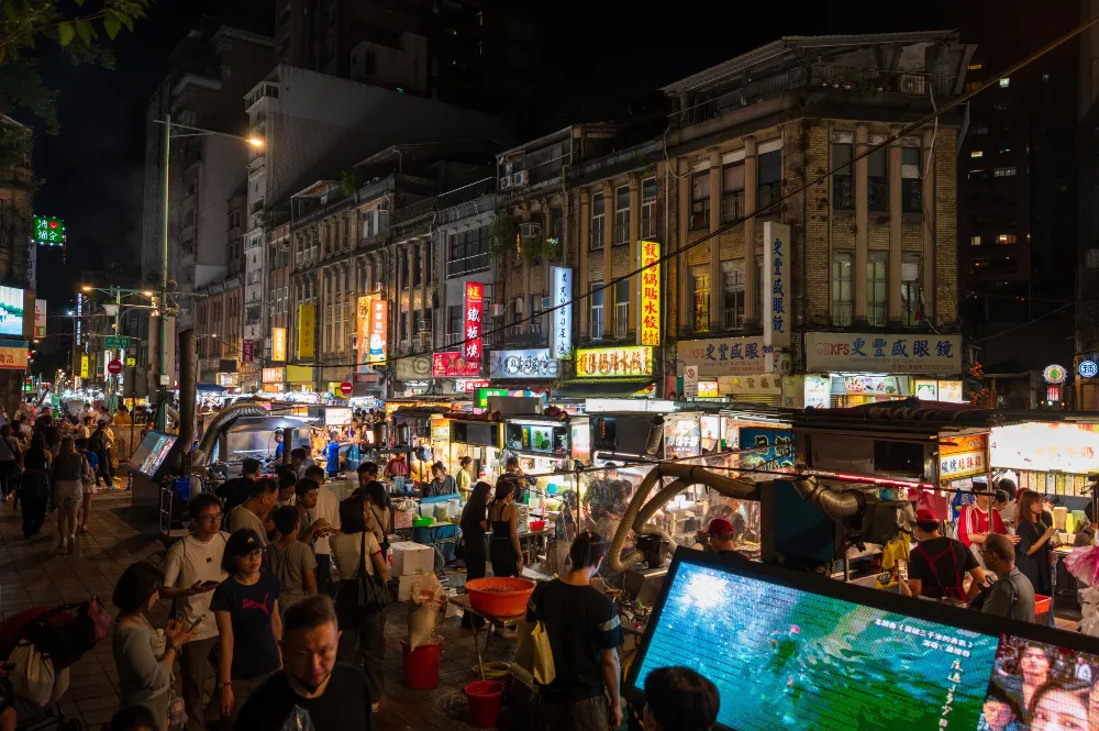 Crowds browse glowing food stalls under neon signs at Ningxia Night Market in Taipei, with old brick buildings above.