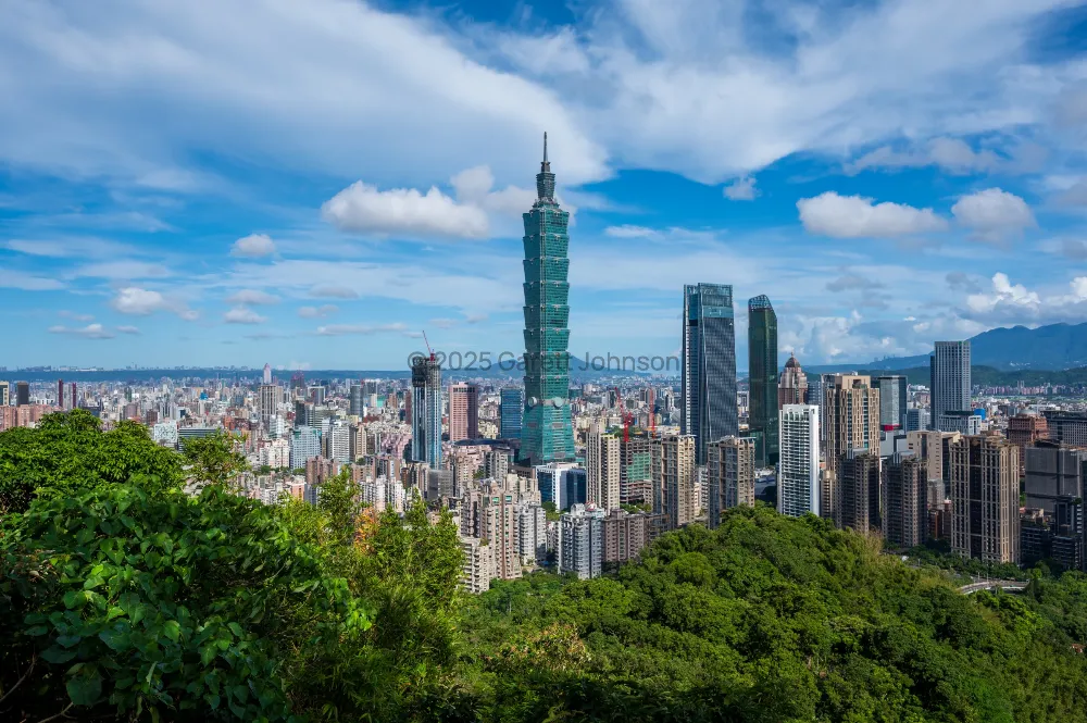 Taipei 101 skyscraper rising above the city skyline, seen from green Elephant Mountain under a blue sky with white clouds.