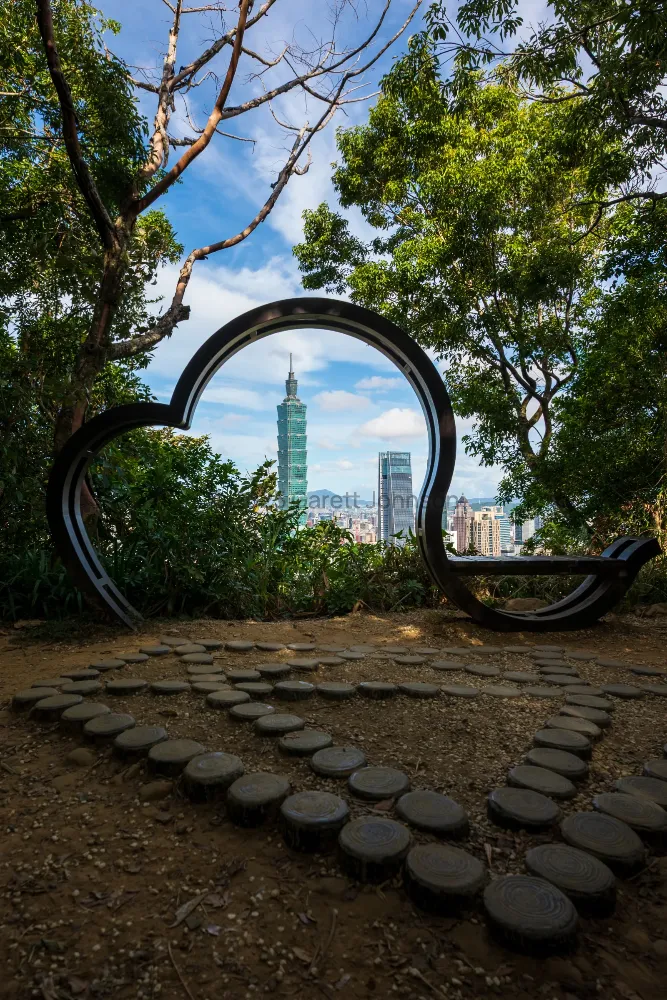 Taipei 101 seen through a curved black circular bench frame on Elephant Mountain, with trees and round stepping stones below.