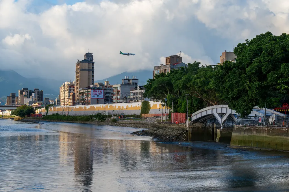 A passenger jet with a green tail descends over Dadaocheng Wharf in Taipei, past riverside buildings, trees and a white arched bridge.