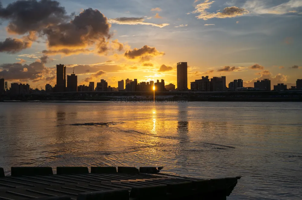 Sun setting behind a city skyline across the river from Dadaocheng Wharf in Taipei, with golden clouds and light on the water.