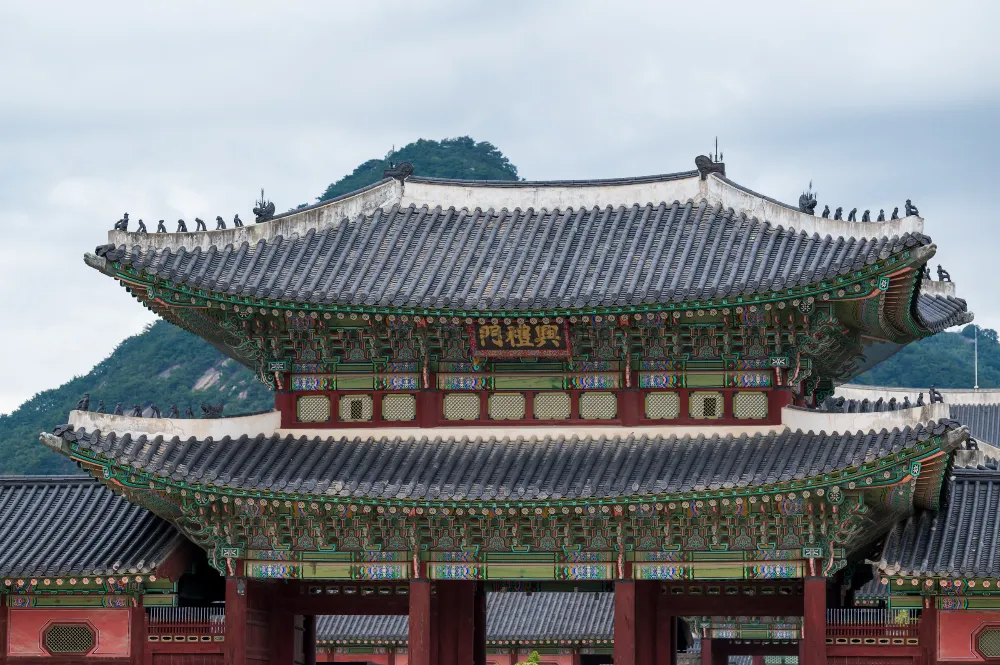 Two-tiered gate at Gyeongbokgung Palace in Seoul, with gray tiled roofs, colorful painted eaves and a forested hill behind.