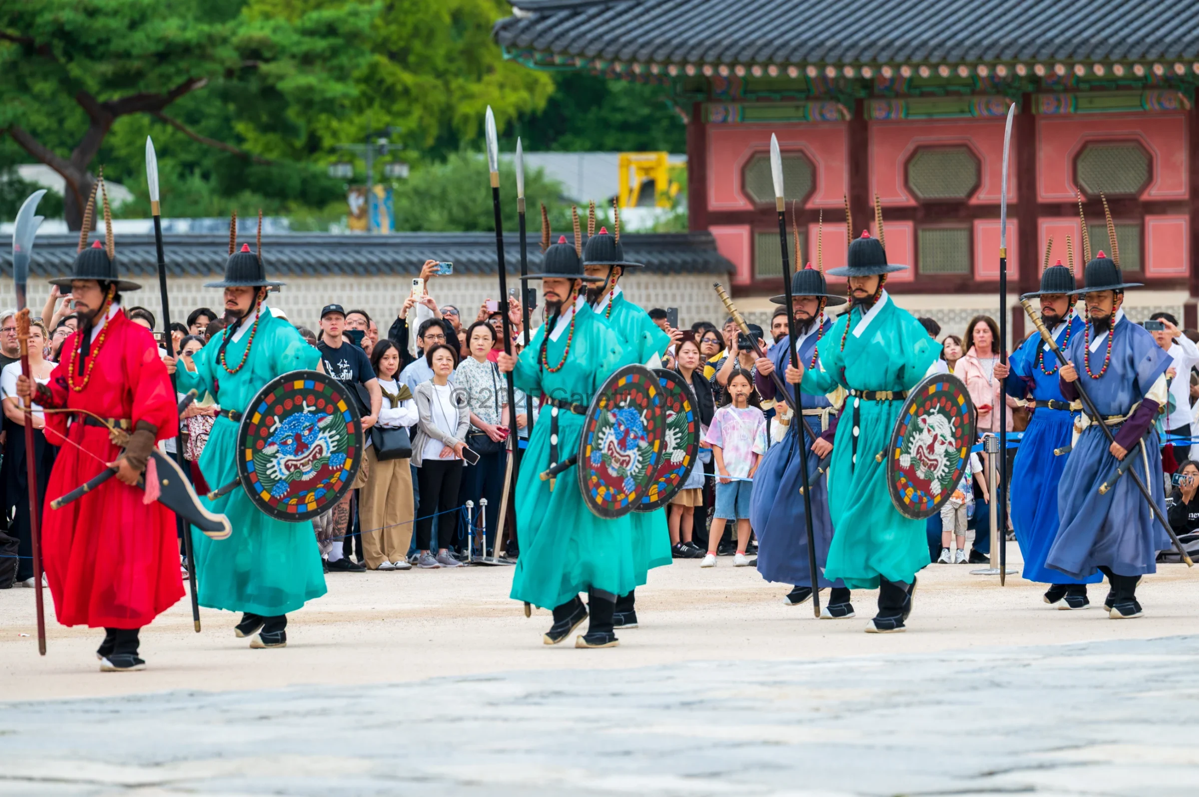 Guards in red, teal and blue traditional robes march with spears and painted shields at Gyeongbokgung Palace as spectators film.
