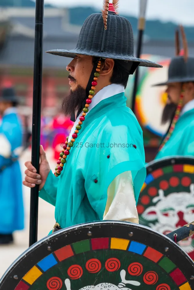 Guard in teal robes and a feathered black hat holds a spear beside a colorful painted shield at Gyeongbokgung Palace, Korea.