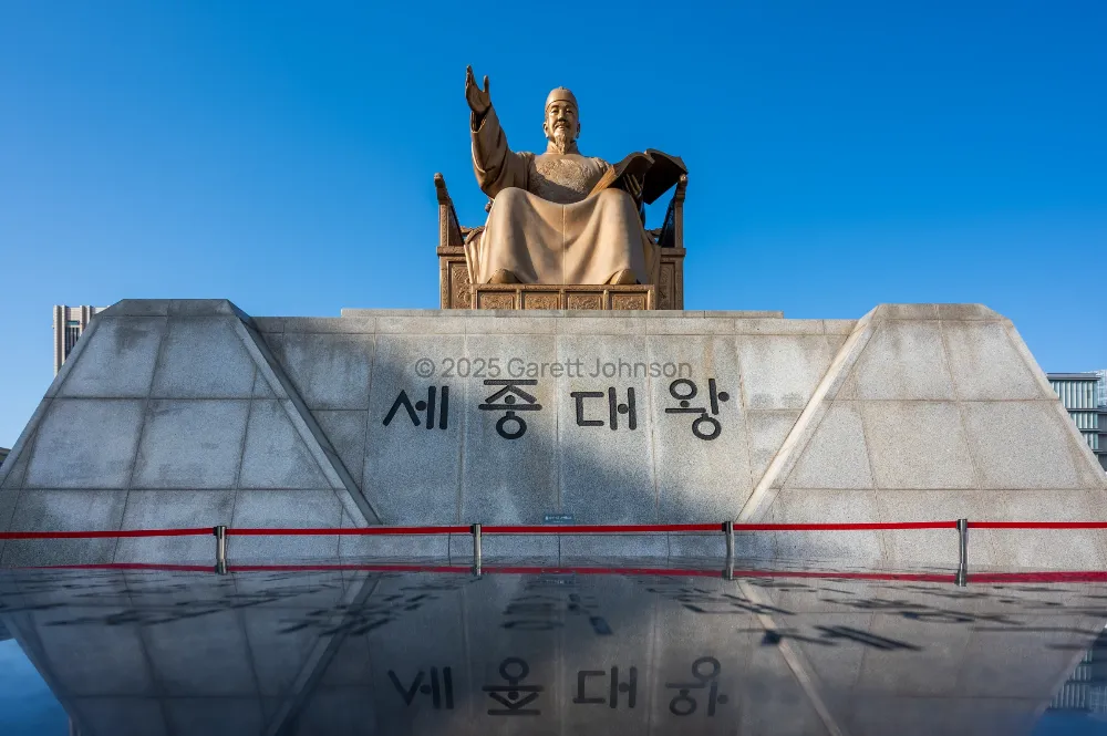 Golden statue of King Sejong seated on a granite pedestal marked in Korean, reflected in water under a clear blue sky in Seoul.
