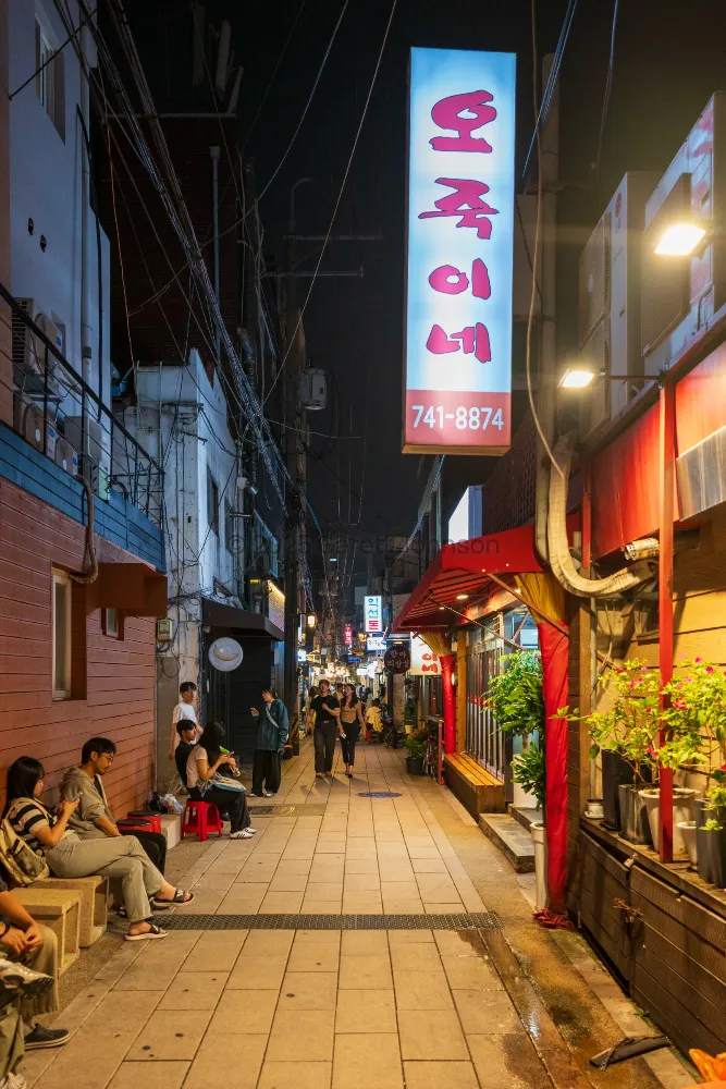 Pedestrians stroll and people sit on benches along a narrow, lantern-lit Seoul alley at night, beneath a glowing Korean sign.