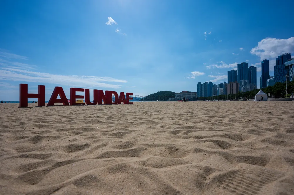 Low view of footprint-covered sand at Haeundae Beach, Busan, with large red HAEUNDAE letters and high-rises under a blue sky.