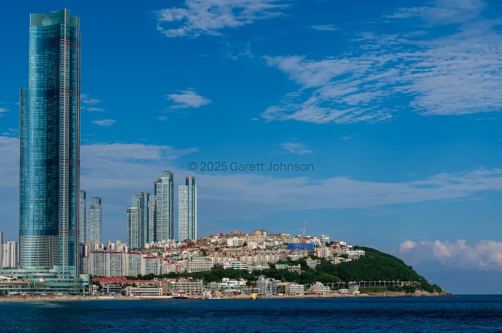 Glass skyscrapers and hillside apartments line Haeundae Beach in Busan beneath a vivid blue sky with wispy clouds.