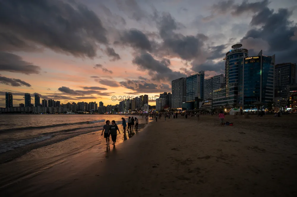 People stroll along the wet sand at Gwangalli Beach in Busan at sunset, with high-rises and dramatic clouds glowing above.