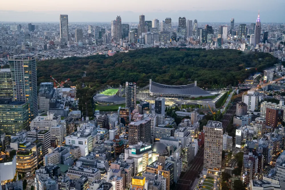 Dusk view from Shibuya Sky over Tokyo, with a dark forested park, a curved-roof stadium and Shinjuku's skyscrapers on the horizon.