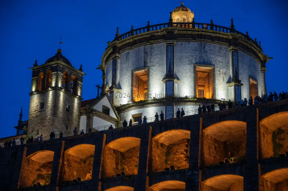 Floodlit round monastery church and bell tower in Porto at dusk, with silhouetted visitors along a wall of glowing stone arches.