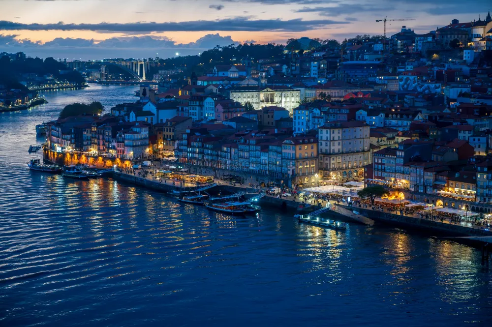 Dusk over Porto's Ribeira district, with lit hillside buildings and riverside cafés reflected in the blue Douro River.