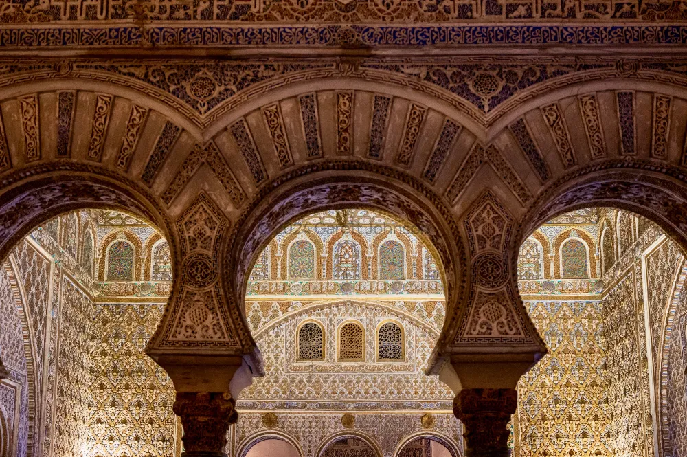 Ornate Moorish horseshoe arches with carved stucco and tilework inside the Real Alcázar in Seville, Spain, in warm light.