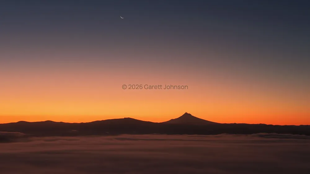 Mt. Hood silhouetted against an orange sunrise sky, with a thin crescent moon above and a fog layer below.
