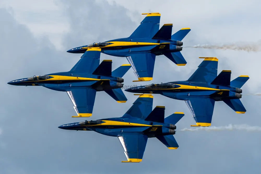 Four blue and yellow Blue Angels jets fly in tight diamond formation against cloudy sky at the Duluth air show, trailing thin smoke.