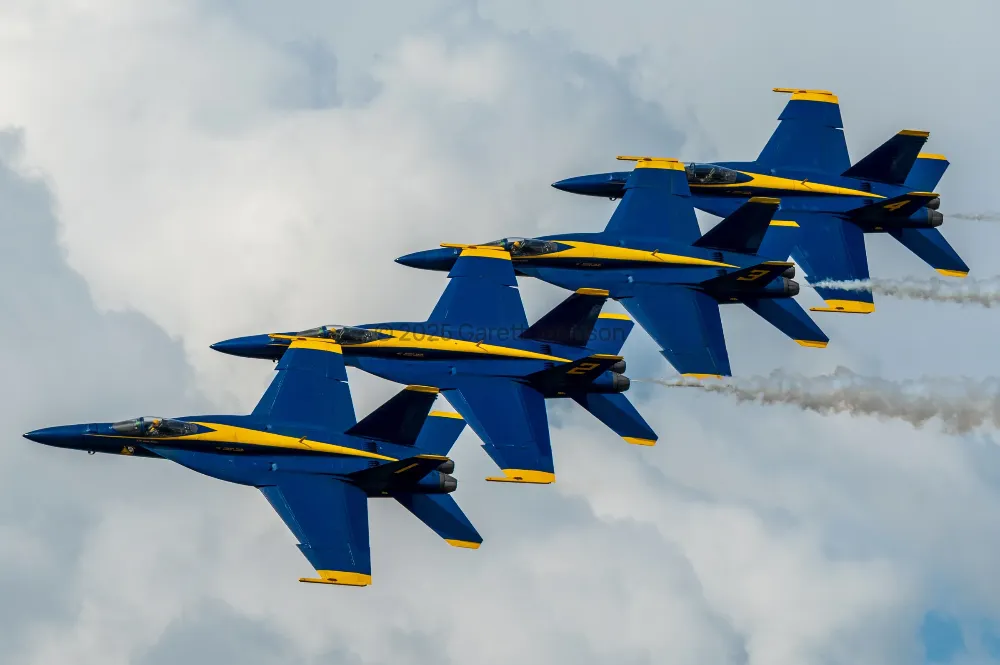 Four Blue Angels jets fly in tight diagonal formation, trailing smoke against billowing clouds at the Duluth Air and Aviation Expo.