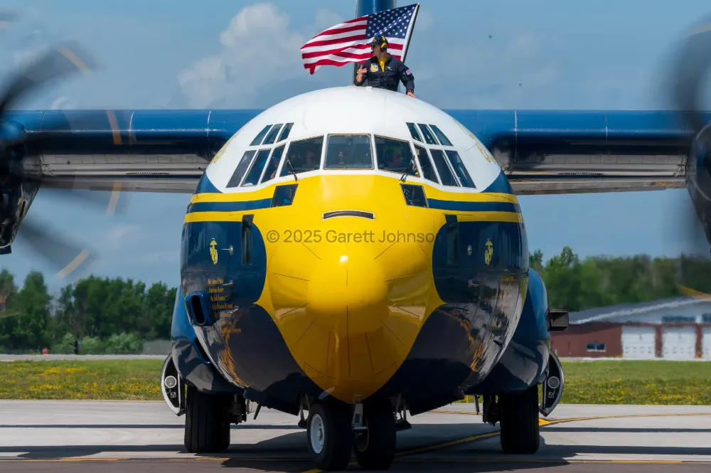 Blue and yellow Fat Albert transport plane taxis head-on as a crew member atop it waves an American flag at the Duluth airshow.