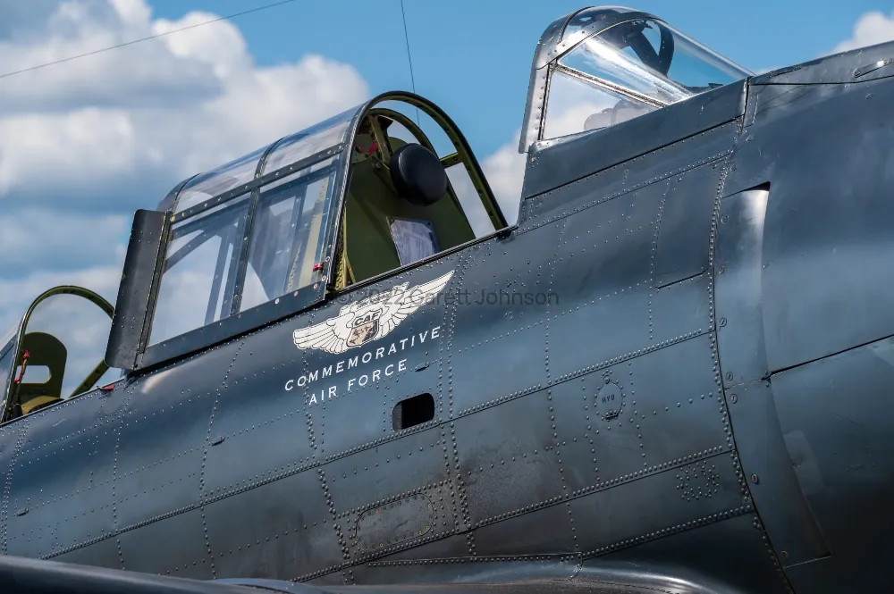 Open cockpits of a riveted dark gray warbird with a Commemorative Air Force winged logo, against a blue sky with clouds.