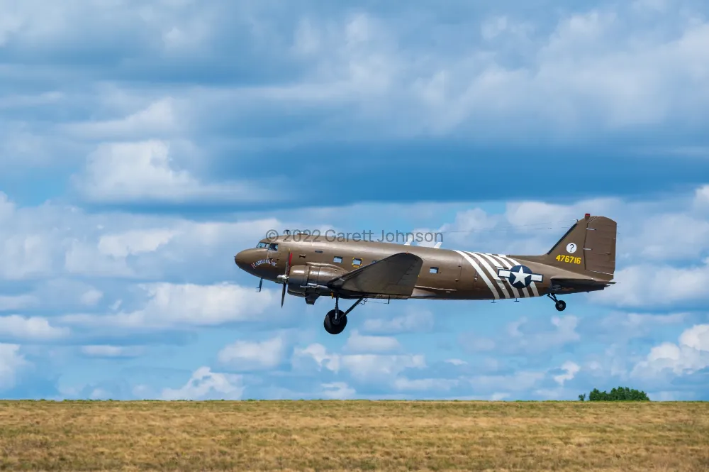 C-47 Skytrain in olive paint with white invasion stripes lifts off over a grassy field under a blue sky with scattered clouds.