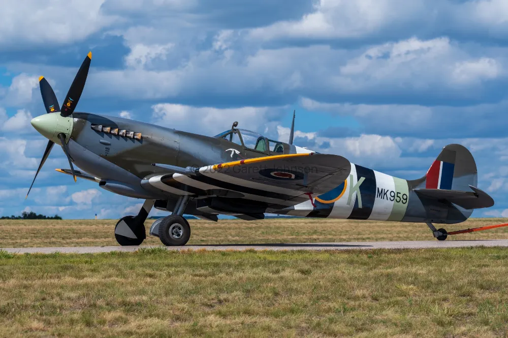 Supermarine Spitfire MK959 with black-and-white invasion stripes parked on the grass at FCM airport under a cloudy blue sky.