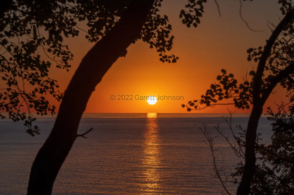 Sunrise over Lake Superior near Tofte, Minnesota, with a glowing orange sky and tree silhouettes framing the sun's reflection.