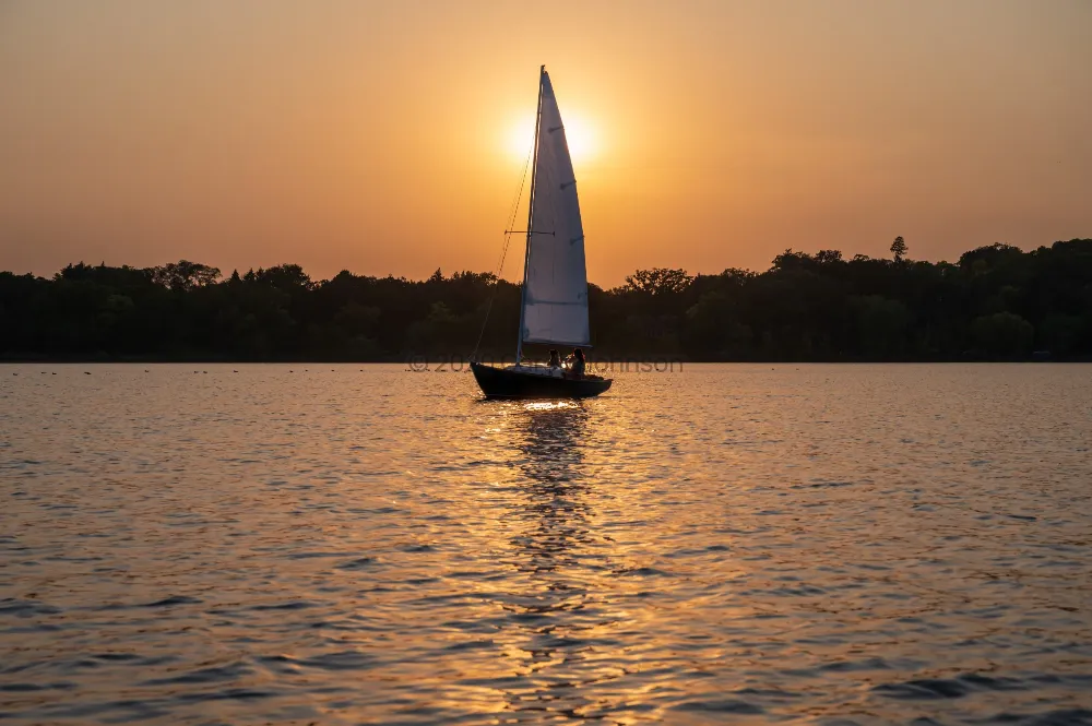 Sailboat silhouetted against a glowing orange sunset on Bde Maka Ska in Minneapolis, with golden reflections on the water.