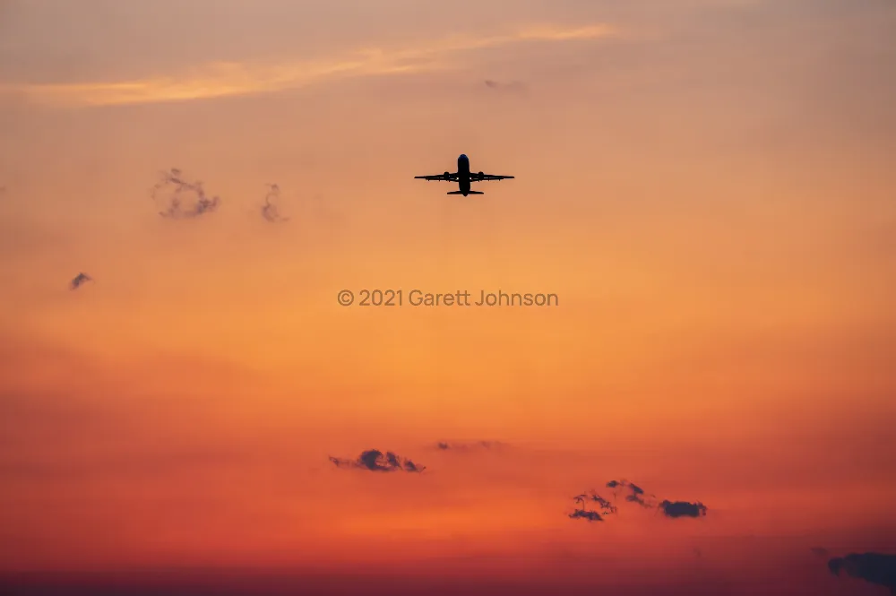 Twin-engine jet silhouetted against an orange and pink sunset sky as it climbs away from MSP, with small scattered clouds.