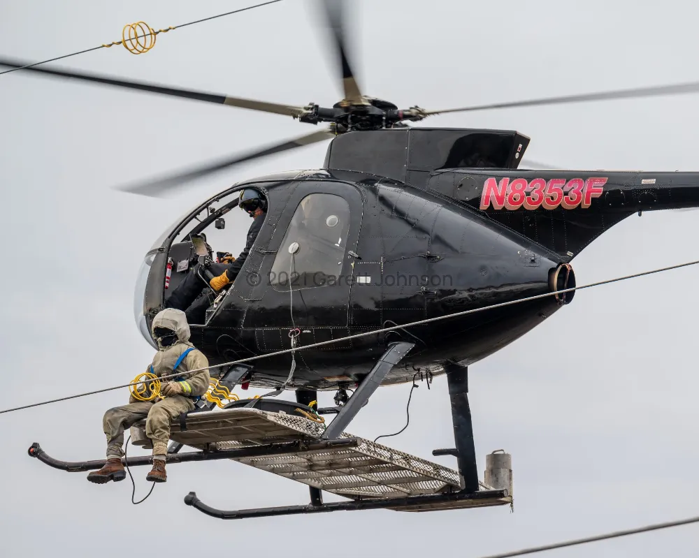 Black Hughes 369D helicopter N8353F hovering beside power lines while a lineman in a protective suit sits on its side platform.