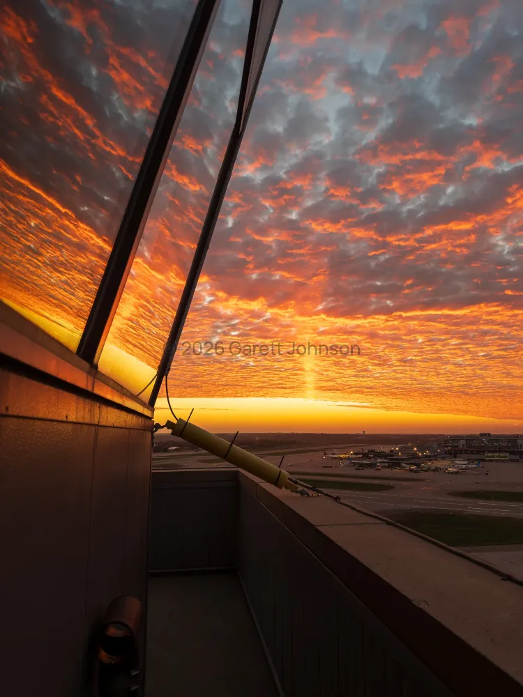 Fiery orange sunset sky of textured clouds reflected in the slanted windows of MSP airport control tower, with the airfield below.