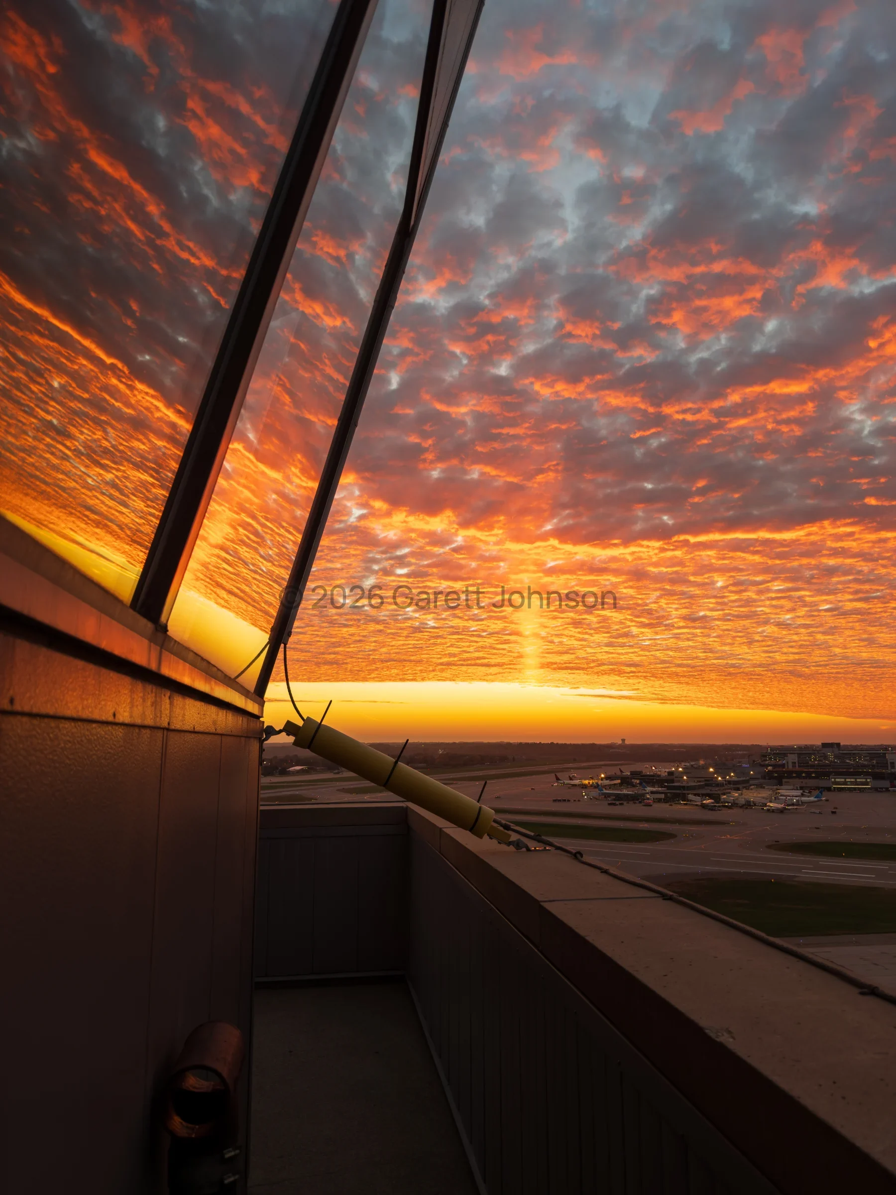 Fiery orange sunset sky of textured clouds reflected in the slanted windows of MSP airport control tower, with the airfield below.