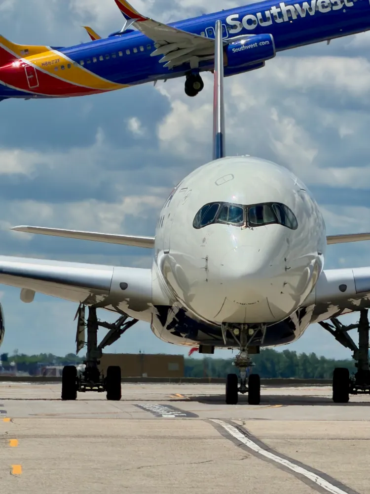 Head-on view of a Delta A350 on the taxiway, its tail aligned with a Southwest 737 passing overhead under cloudy skies.
